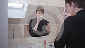 Beautiful brunette mature woman looking at big mirror and smelling face creams. Stylish Caucasian lady applying makeup