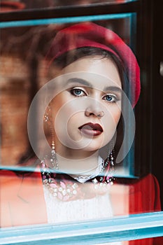 Beautiful brunette model with blue eyes in a red beret is looking through the glass at the camera. Skin retouching