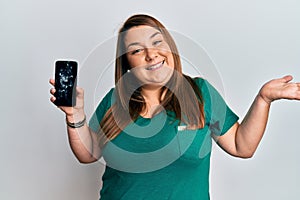 Beautiful brunette plus size woman holding broken smartphone showing cracked screen celebrating achievement with happy smile and