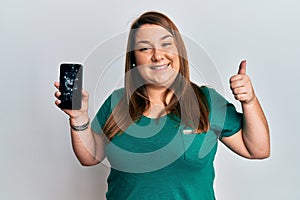 Beautiful brunette plus size woman holding broken smartphone showing cracked screen smiling happy and positive, thumb up doing