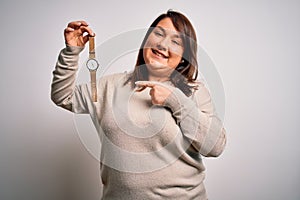 Beautiful brunette plus size woman holding wristwatch over isolated white background very happy pointing with hand and finger