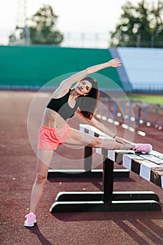 Beautiful brunette runner woman doing stretching leaning her leg on barrier for running stretching before workout - outdoor shot.