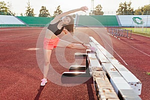 Beautiful brunette runner woman doing stretching leaning her leg on barrier for running stretching before workout - outdoor shot.