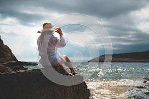 Beautiful brunette seen from behind sitting on a rock on the shores of adriatic sea, cloudy day during the summer of 2021. Waves