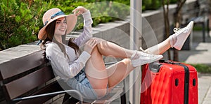 Beautiful brunette in shorts and a hat sits on a bench stretched out legs on a large red suitcase. Caucasian woman