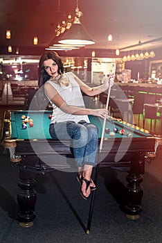 Beautiful brunette sitting on billiard table with cue