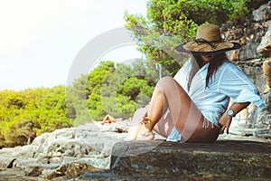 Beautiful brunette sitting on a rock on a beach in Croatia. Wearing a hay hat and a white shirt, warm sunny day, happy smile while