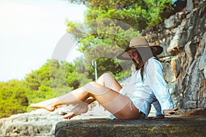 Beautiful brunette sitting on a rock on a beach in Croatia. Wearing a hay hat and a white shirt, warm sunny day, happy smile while