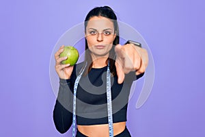 Beautiful brunette sporty woman using tape measure eating apple fruit over purple background pointing with finger to the camera