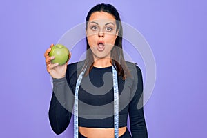 Beautiful brunette sporty woman using tape measure eating apple fruit over purple background scared and amazed with open mouth for