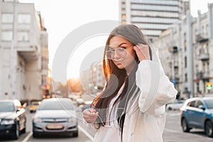 Beautiful brunette in a white jacket and glasses listens to music in the headphones at the background of a city landscape in the