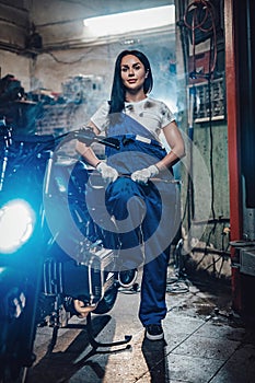 Beautiful brunette woman in blue overalls posing next to a custom bobber in garage or workshop