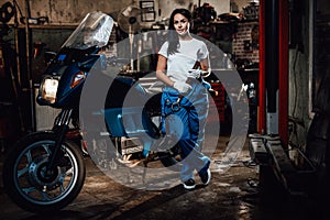 Beautiful brunette woman in blue overalls posing next to a sportbike in garage or workshop