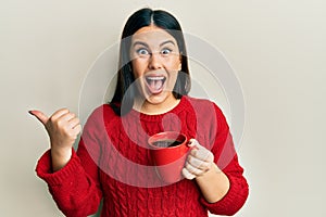 Beautiful brunette woman drinking a cup of black coffee pointing thumb up to the side smiling happy with open mouth