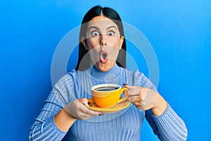 Beautiful brunette woman drinking a yellow cup of black coffee afraid and shocked with surprise and amazed expression, fear and
