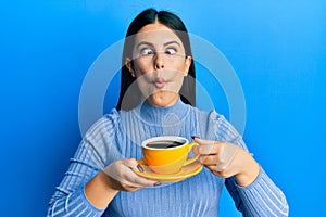 Beautiful brunette woman drinking a yellow cup of black coffee making fish face with mouth and squinting eyes, crazy and comical