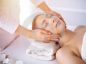 Beautiful brunette woman enjoying facial massage with closed eyes in sunny spa center . Relaxing treatment and cosmetic