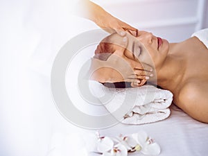 Beautiful brunette woman enjoying facial massage with closed eyes in sunny spa center . Relaxing treatment and cosmetic