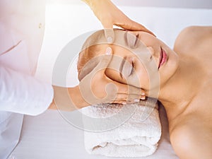 Beautiful brunette woman enjoying facial massage with closed eyes in sunny spa center . Relaxing treatment and cosmetic