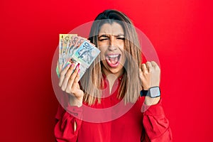 Beautiful brunette woman holding australian dollars angry and mad raising fist frustrated and furious while shouting with anger