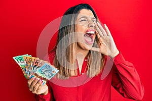 Beautiful brunette woman holding australian dollars shouting and screaming loud to side with hand on mouth