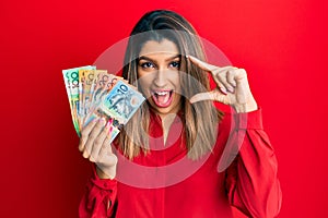 Beautiful brunette woman holding australian dollars smiling and confident gesturing with hand doing small size sign with fingers