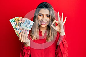 Beautiful brunette woman holding australian dollars smiling positive doing ok sign with hand and fingers