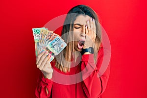 Beautiful brunette woman holding australian dollars yawning tired covering half face, eye and mouth with hand