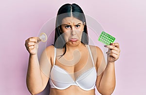 Beautiful brunette woman holding condom and birth control pills depressed and worry for distress, crying angry and afraid