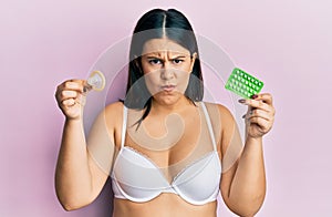 Beautiful brunette woman holding condom and birth control pills skeptic and nervous, frowning upset because of problem