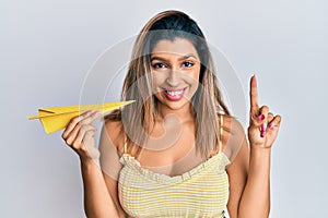 Beautiful brunette woman holding paper airplane smiling with an idea or question pointing finger with happy face, number one