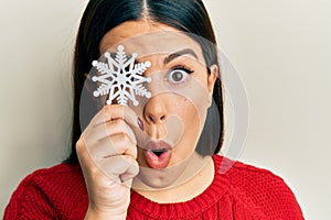 Beautiful brunette woman holding snowflake over eye scared and amazed with open mouth for surprise, disbelief face