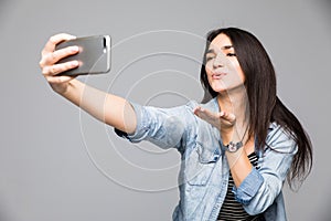 Beautiful brunette woman making a selfie blowing a kiss holding the smartphone isolated on a gray background