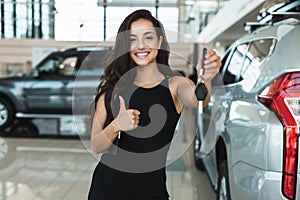 Beautiful brunette woman manager smiling holding car keys in her hand standing in dealership center near brand new SUV photo