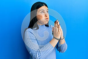 Beautiful brunette woman praying holding catholic rosary puffing cheeks with funny face