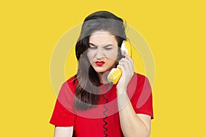 A beautiful brunette woman with red lipstick speaks on an old yellow telephone with a cord. Woman is unhappy, does not want to