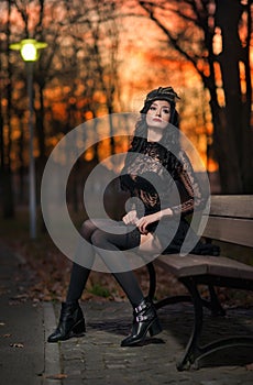 Beautiful brunette woman sitting outside in park with dramatic sky on background. Fashionable female with cap resting on bench