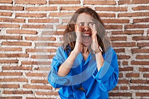 Beautiful brunette woman standing over bricks wall shouting angry out loud with hands over mouth