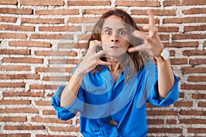 Beautiful brunette woman standing over bricks wall shouting frustrated with rage, hands trying to strangle, yelling mad