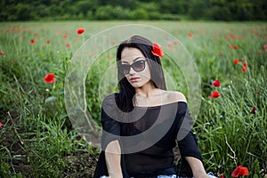 Beautiful brunette woman in sunglasses and stylish clothes sitting among on red poppies