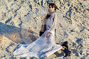 Beautiful brunette woman in translucent beach cover up with leopard fur coat and black boots posing on sandy beach at sunset.