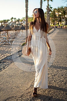 Beautiful brunette woman walking on sunset beach in fashionable maxi dress, relaxing on luxury tropical summer holidays