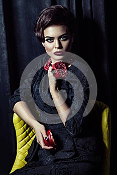 Beautiful brunette woman wearing black evening dress posing with two pomegranates in hands while sitting on yellow chair