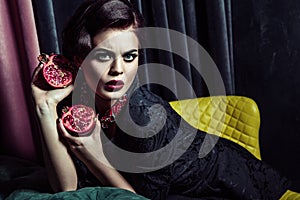 Beautiful brunette woman wearing black evening dress posing with two pomegranates in hands while sitting on yellow chair