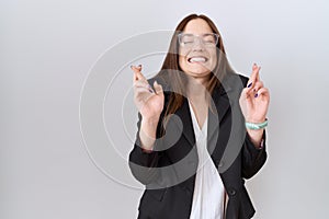 Beautiful brunette woman wearing business jacket and glasses gesturing finger crossed smiling with hope and eyes closed