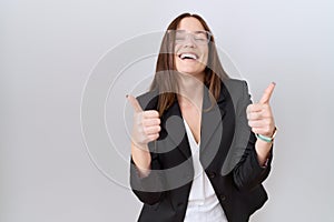 Beautiful brunette woman wearing business jacket and glasses success sign doing positive gesture with hand, thumbs up smiling and