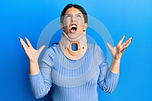 Beautiful brunette woman wearing cervical collar crazy and mad shouting and yelling with aggressive expression and arms raised