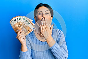 Beautiful brunette woman wearing cervical collar holding insurance money covering mouth with hand, shocked and afraid for mistake