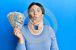 Beautiful brunette woman wearing cervical collar holding insurance money scared and amazed with open mouth for surprise, disbelief