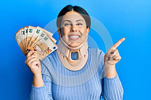Beautiful brunette woman wearing cervical collar holding insurance money smiling happy pointing with hand and finger to the side